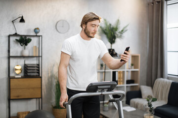 Naklejka na ściany i meble Front view of caucasian man in sports clothes using smart phone for video call doing cardio training on treadmill at home or gym. Concept of sport, health care, action, remote leisure.