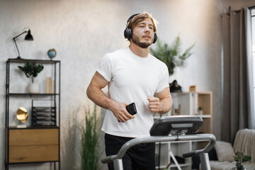 Close up portrait of handsome muscular man in white t-shirt having workout on his electric treadmill, running and listening favorite music using smartphone and headphones.