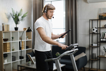 Fototapeta na wymiar Side view of caucasian man in sport clothes using headset working tablet computer while doing cardio training on treadmill at morning at home gym. Concept of sport, health care, business.