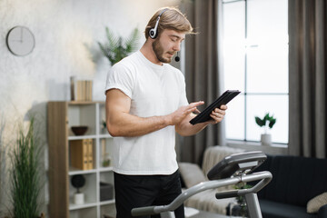 Fototapeta na wymiar Smiling muscular sporty businessman in headset working remote from home using digital tablet, while having cardio workout, running on treadmill.