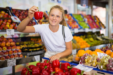 Young positive girl choosing fresh bell pepper at grocery section of supermarket