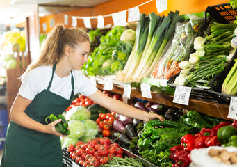 Positive young female seller in uniform holding green pepper in grocery shop