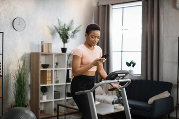 Fototapeta na wymiar Portrait of active african woman in sportswear using smartphone, running using treadmill at home at morning. Coronavirus Covid 19 social distance. Home workout, stay at home, remote leisure concept.