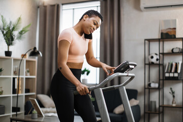 Side view of african sporty woman in pink t-shirt working out in treadmill while having phone call...