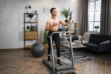 Fototapeta na wymiar Healthy young sports woman using phone having video conference while working out, running, doing cardio training on treadmill in morning time, indoor on background of modern apartment