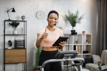 Fototapeta na wymiar Portrait of active and dynamic young pretty businesswoman with headset and tablet doing sport fitness at home running on treadmill indoor at night showing thumb up. Tone your body. Perfect shape.