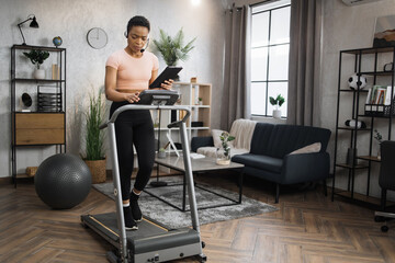 Fototapeta premium Side view of african woman in sport clothes using headset working on tablet computer while doing cardio training on treadmill at morning at home gym. Concept of sport, health care, business.