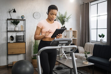 Fototapeta premium Confident muscular sporty businesswoman in headset working remote from home using digital tablet, while having cardio workout, running on treadmill.