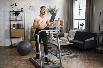 Fototapeta na wymiar Full length photo of young active african business woman in sportswear and headset training at home working with tablet, doing cardio exercise on treadmill. Concept of sport, business.