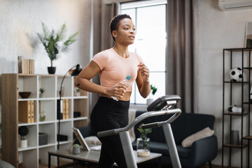 Fototapeta premium Attractive young sports african american woman holding bottle with water working out, running, doing cardio training on treadmill, indoor on background of modern apartment.