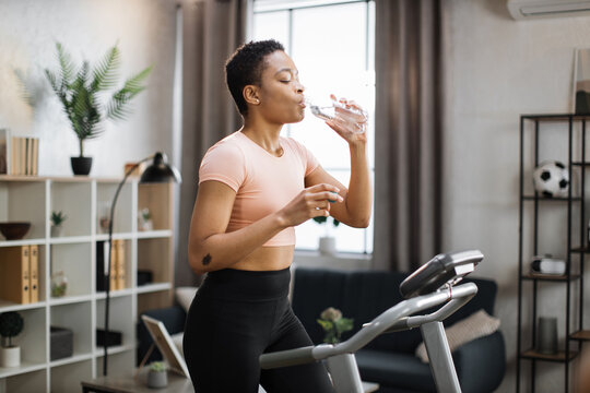Pretty young sports african woman drinking water from bottle doing fitness exercise, running on treadmill . Athletic and muscular female having workout indoor, at modern apartment at home.