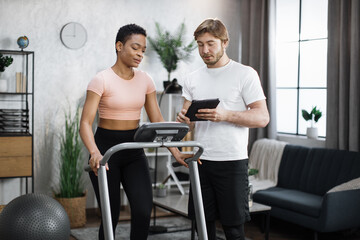 Fototapeta na wymiar Fitness caucasian man trainer using tablet to show online lesson for african american woman training on treadmill at modern home. Two sporty people engaged in remote workout .