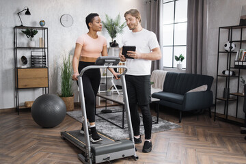 Obraz premium Portrait of african american female wearing sportswear working out using treadmill and speak to caucasian man trainer. Home fitness workout two people training on smart stationary running track.