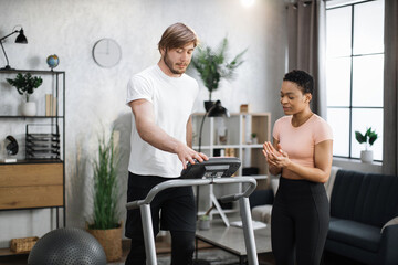 Fit african woman coach using control panel while training young caucasian man to run on treadmill. Attractive sporty fit people using running track and choosing mode for sports training.