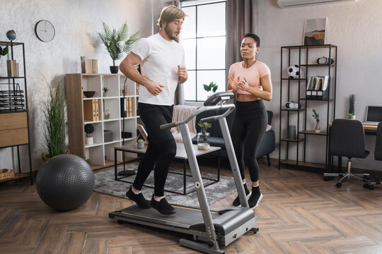 Fit African Woman Coach Using Control Panel While Training Young Caucasian Man To Run On Treadmill. Attractive Sporty Fit People Using Running Track And Choosing Mode For Sports Training.