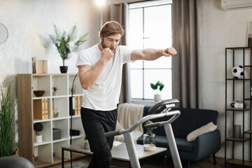 Obraz na płótnie Canvas Strong young caucasian man doing boxing punches while training indoors. Professional bearded muscle sportsman boxing and exercising warm-up during home workout at treadmill.