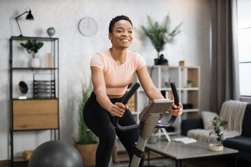 Smiling beautiful african american sports woman in sportswear cycling bike at home on background of...