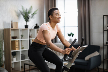 Fototapeta na wymiar Smiling beautiful african american sports woman in sportswear cycling bike at home on background of light living room. Cardio training, exercising legs, cardio workout indoors.