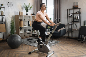African smiling young sportswoman doing cardio on stationary bike. Attractive female working out on fitness in the morning at home. Woman training on exercise bike indoors.