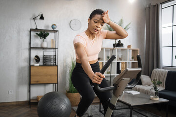 Fototapeta na wymiar Tired african american sporty woman in pink t-shirt leaned her hand on exercise bike and another wiping sweat from forehead while working out in morning at living room at home.