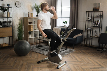 Thirsty handsome young sports bearded caucasian man in sportswear drinking fresh water while cycling bike at home. Cardio training, exercising legs, cardio workout indoors.