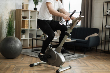 Cropped view of young male with water bottle wearing sportswear using exercise bike. Home fitness workout sporty man training on smart stationary bike indoors. Young caucasian guy athlete.