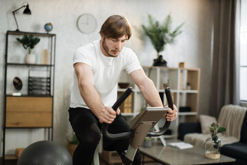 Naklejka na ściany i meble Home fitness workout, young caucasian man athlete training on smart stationary bike indoors watching on screen connected online to live streaming subscription service for biking exercise.