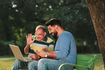 Young indian man showing bank card and laptop how to uses to his father at park