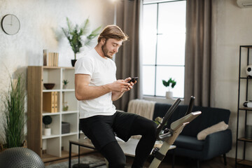 Portrait of focused young male wearing sportswear using exercise bike typing message on phone. Home fitness workout sporty man training on smart stationary bike indoors.