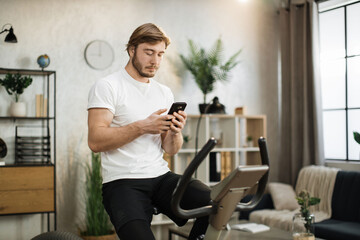 Portrait of focused young male wearing sportswear using exercise bike typing message on phone. Home...