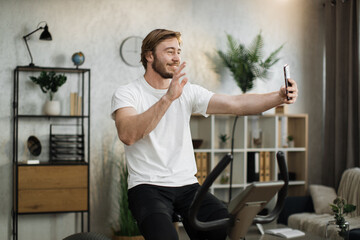Handsome young sports bearded caucasian man in sportswear having online video call while cycling bike at home. Cardio training, exercising legs, cardio workout indoors.