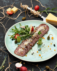 Baked meat cut into slices with vegetables and Parmesan. Plate with dish on dark background. Cheese knife. 