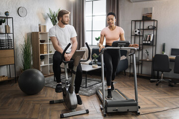 Portrait of focused caucasian male and african female wearing sportswear using exercise bike and treadmill. Home fitness workout sporty people training on exercise machines indoors.