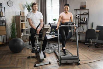 Portrait of focused caucasian male and african female wearing sportswear using exercise bike and treadmill. Home fitness workout sporty people training on exercise machines indoors.