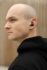 Fototapeta premium Young bald man with freckles on face. Guy wearing in black hoodie and with wireless headphones. Portrait in profile. Wooden background.