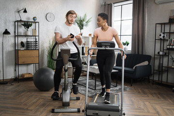 Happy smiling couple doing cardio on stationary bike and treadmill using smart phone to watch online video lessons how to sole weight. Attractive female and male working out on fitness at home.