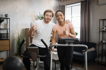Fototapeta na wymiar African woman running on treadmill and caucasian man training on exercise bike. Young couple doing cardio on stationary bike and treadmill. Attractive female and male working out at home.