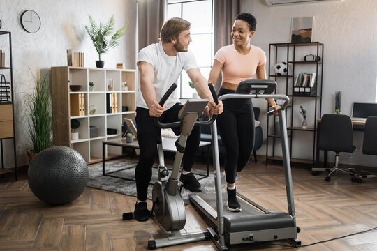 Full length portrait of focused caucasian male and african female wearing sportswear using exercise bike and treadmill. Home fitness workout sporty people training on exercise machines indoors