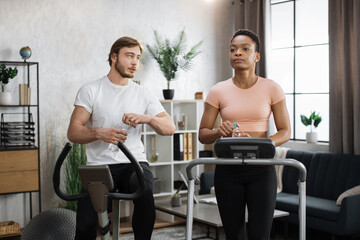 Fototapeta na wymiar Portrait of young caucasian male and african female wearing sportswear using exercise bike and treadmill drinking water. Home fitness workout sporty people training on exercise machines