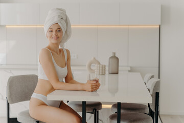 Relaxed beautiful slender woman sitting in kitchen after taking shower with glass of water