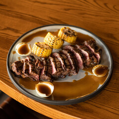 Grilled tender medium rare flank steak sliced and served with roast corn on cob and sauce on plate. Plate with dish on wooden table. View from above. 