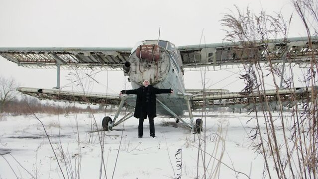 An old broken aircraft AN 2 in the middle of a snowy landscape. A man in a dark coat and a white cap spread his hands parallel to his wings.