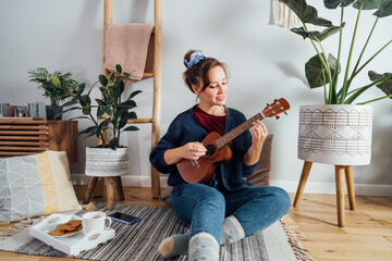 Young woman playing ukulele guitar while relaxing on floor cushion in modern scandinavian home interior.Musical Hobby, home fun concept. Resolution to learn new skills. Selective focus,