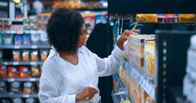 African American young woman shopper, in white casual shirt and blue denim jeans, attentively reading the content on label while shopping for cereals in the grocery store