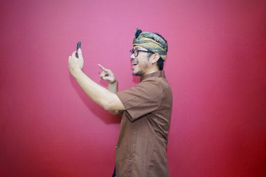 A portrait of a happy Balinese man is smiling and holding his smartphone wearing udeng and and traditional clothes isolated by a red background. Concept photo on a video call.