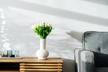 Scandinavian home interior with spring bouquet of white tulip flowers in ceramic vase, tray with candles standing on a wooden cabinet. Minimalist design with gray sofa and white wall. Springtime.