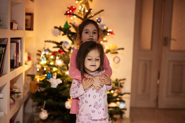 Two sisters together near Christmas tree at evening home.