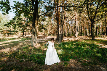 Young beautiful girl in elegant dress is standing and holding hand bouquet of white flowers and greens with ribbon at nature. The bride holds a wedding bouquet outdoors.