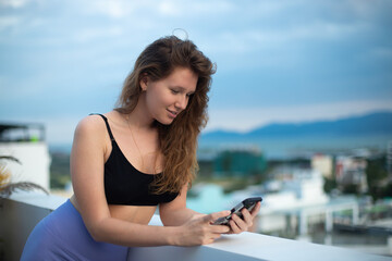 Fototapeta na wymiar happy young girl uses her phone on the roof of a house, balcony, city background, a woman taking selfie at summer, have a video chat, smiling have fun, photo of herself on mobile phone, smartphone 