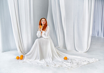 beautiful red-head girl in white dress with long curly hair holding oranges  and sitting in white studio with curtains